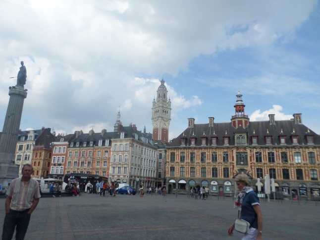 A bit of Grand Place, featuring: 1) Colonne de la Déesse [Column of the Goddess] to the left (a memorial to the 1792 siege of Lille by Austrian forces) 2) Various shops 3) The belfry of the beautiful post office, centre-back 4) La Vieille Bourse [The Old Stock Exchange] 5) Photobombing Lillois, left and right foreground